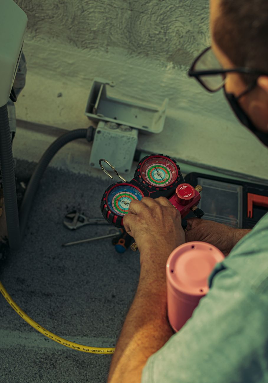 Close-up of a technician's hands holding a red and blue refrigerant manifold gauge set next to an outdoor air conditioning unit, with a yellow charging hose on the ground
