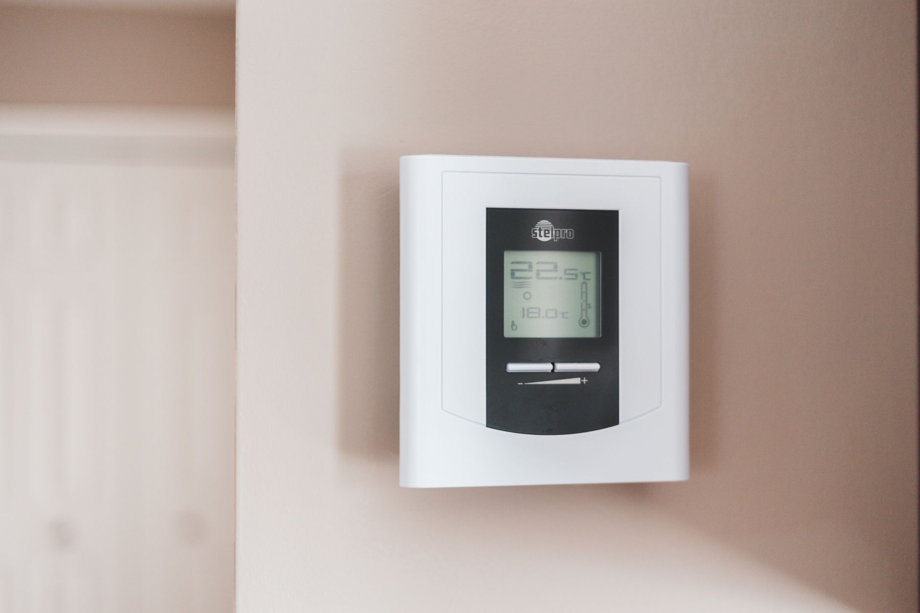 A white digital wall thermostat with a gray display panel showing room and set temperatures, mounted on a pale pink wall