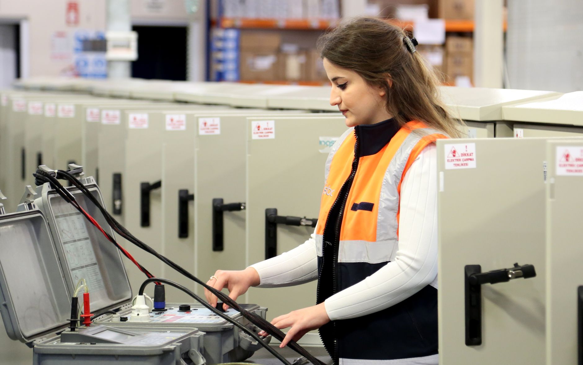 Technician in an orange high-visibility vest running tests with a portable electrical test instrument in front of a row of beige electrical control cabinets