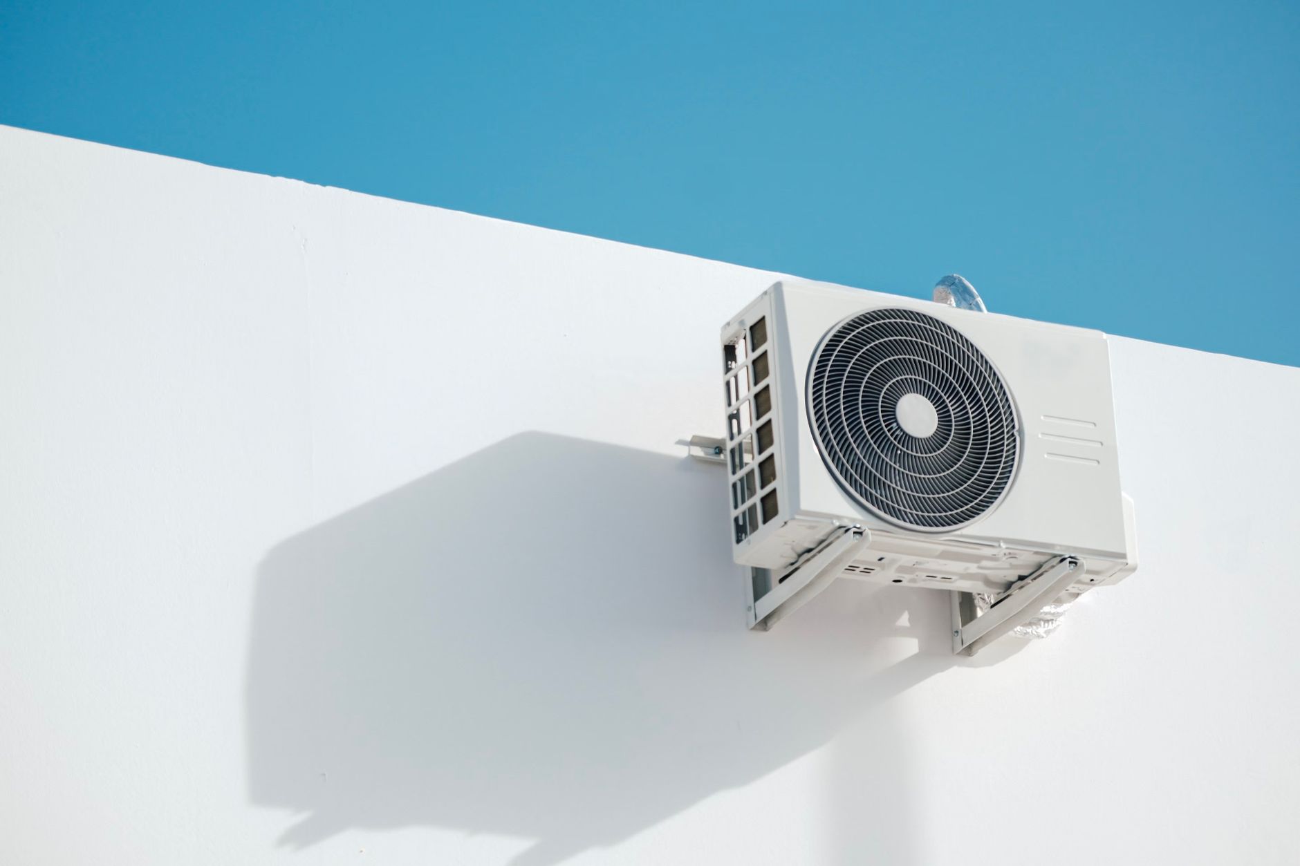 The outdoor unit of a ductless split system mounted on brackets high on a white wall under a blue sky