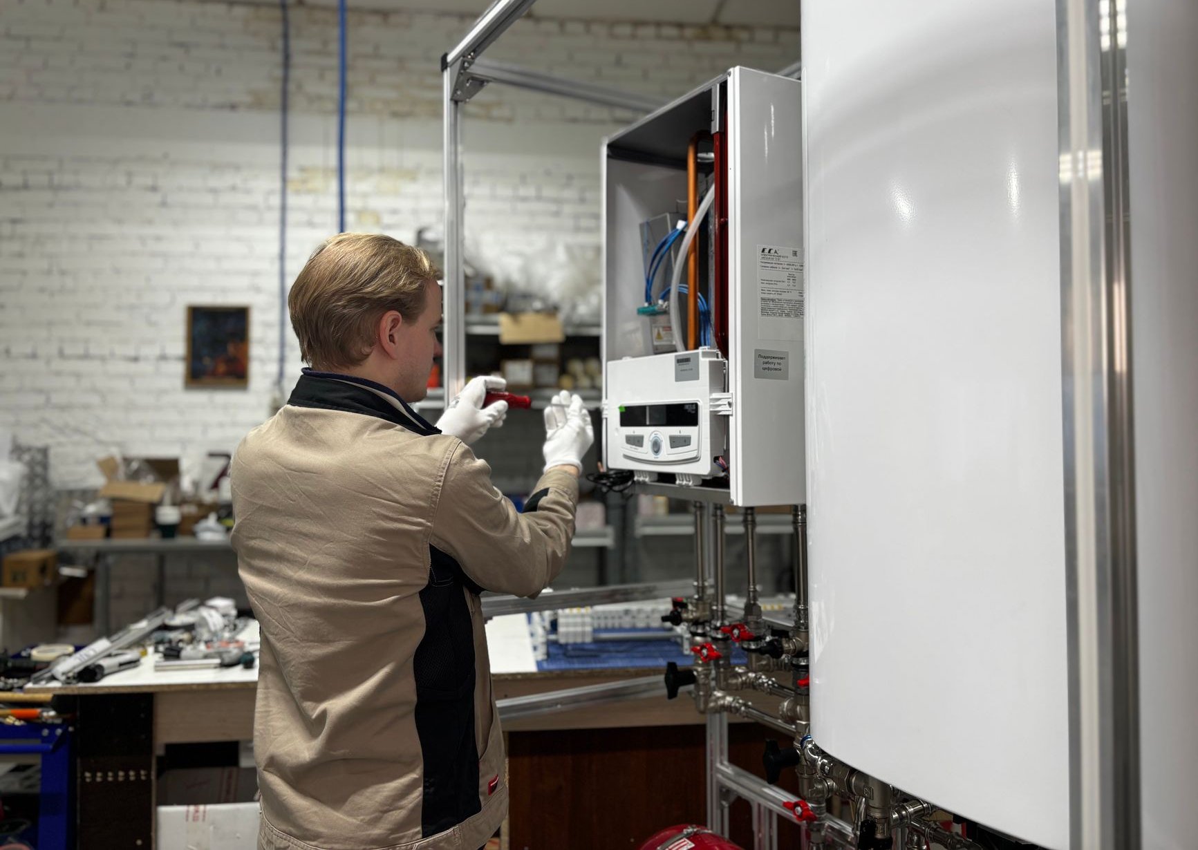 A young technician in a work jacket and white gloves examines the open control panel of a wall-mounted gas boiler on a training rig in a workshop