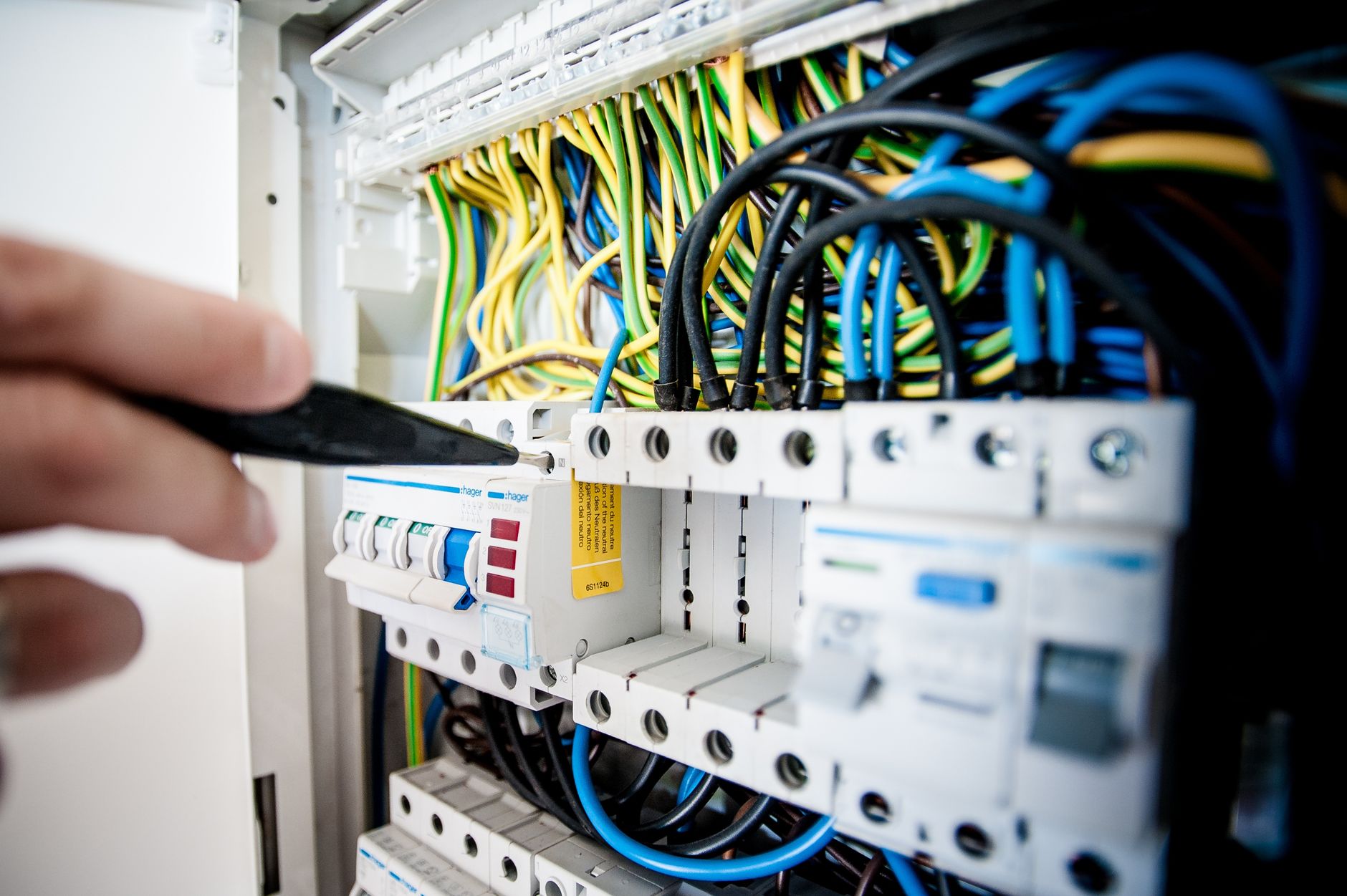 A hand pointing a screwdriver at circuit breakers in an open electrical panel full of blue, black and yellow-green wires