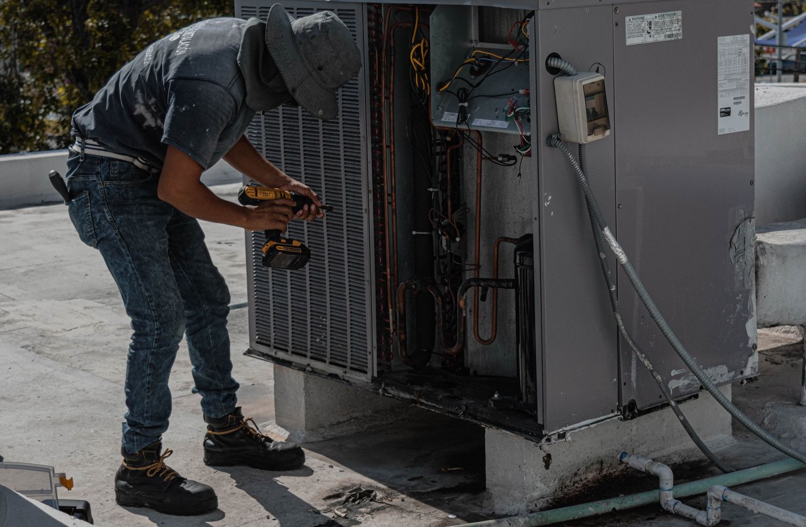 A technician in a bucket hat and work boots uses a cordless drill on a rooftop air conditioning unit with its side panel removed, showing copper tubing and wiring