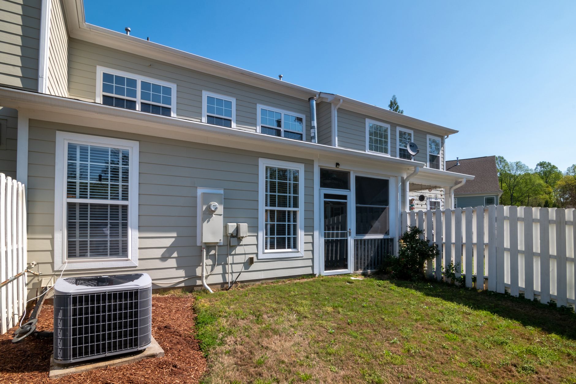 Back of a gray two-story townhouse with a residential air conditioning condenser on a pad in a mulch bed beside a white fence