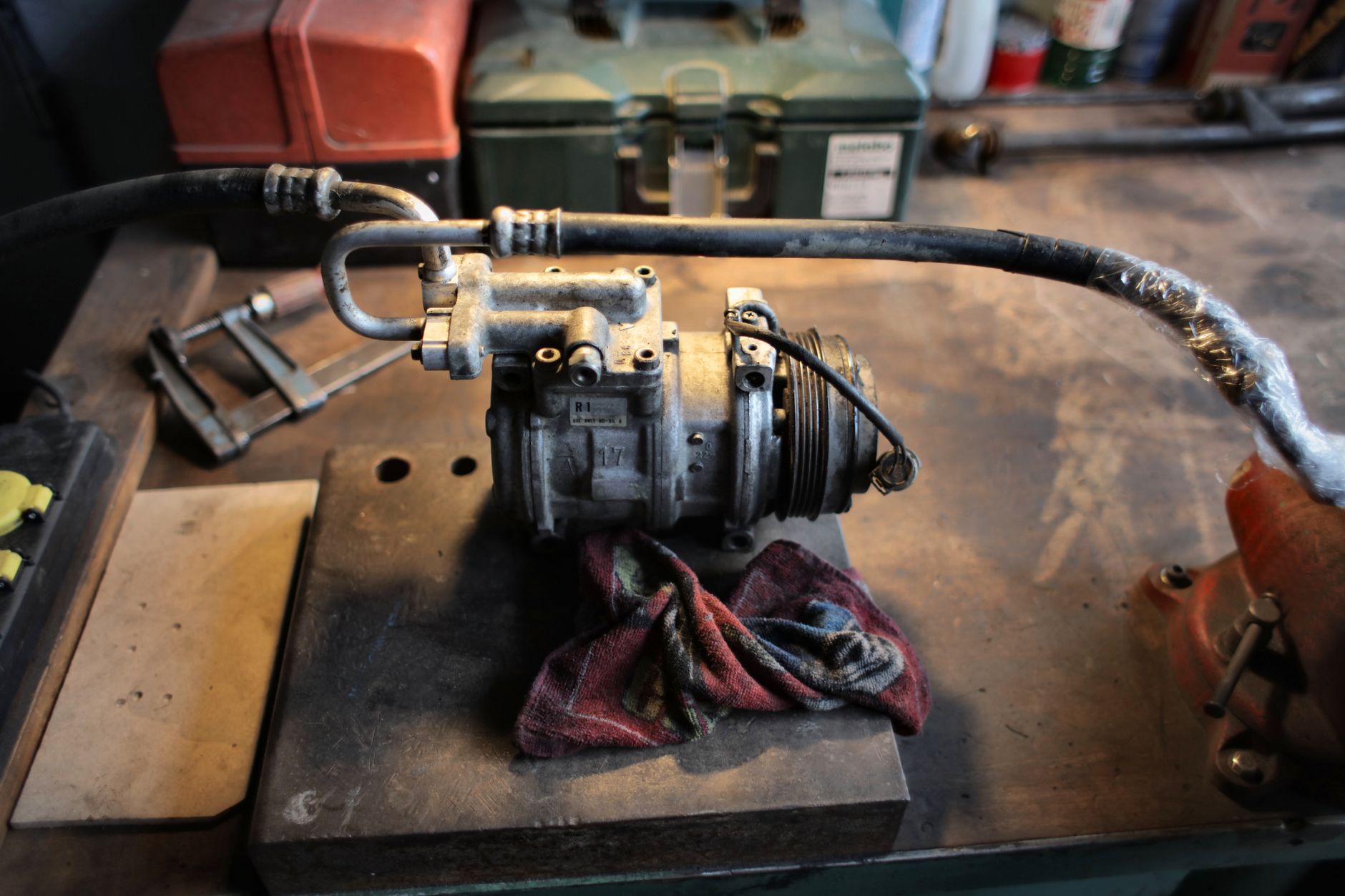 A used air conditioning compressor with its refrigerant line attached sits on a steel workbench beside a red rag, a clamp and toolboxes
