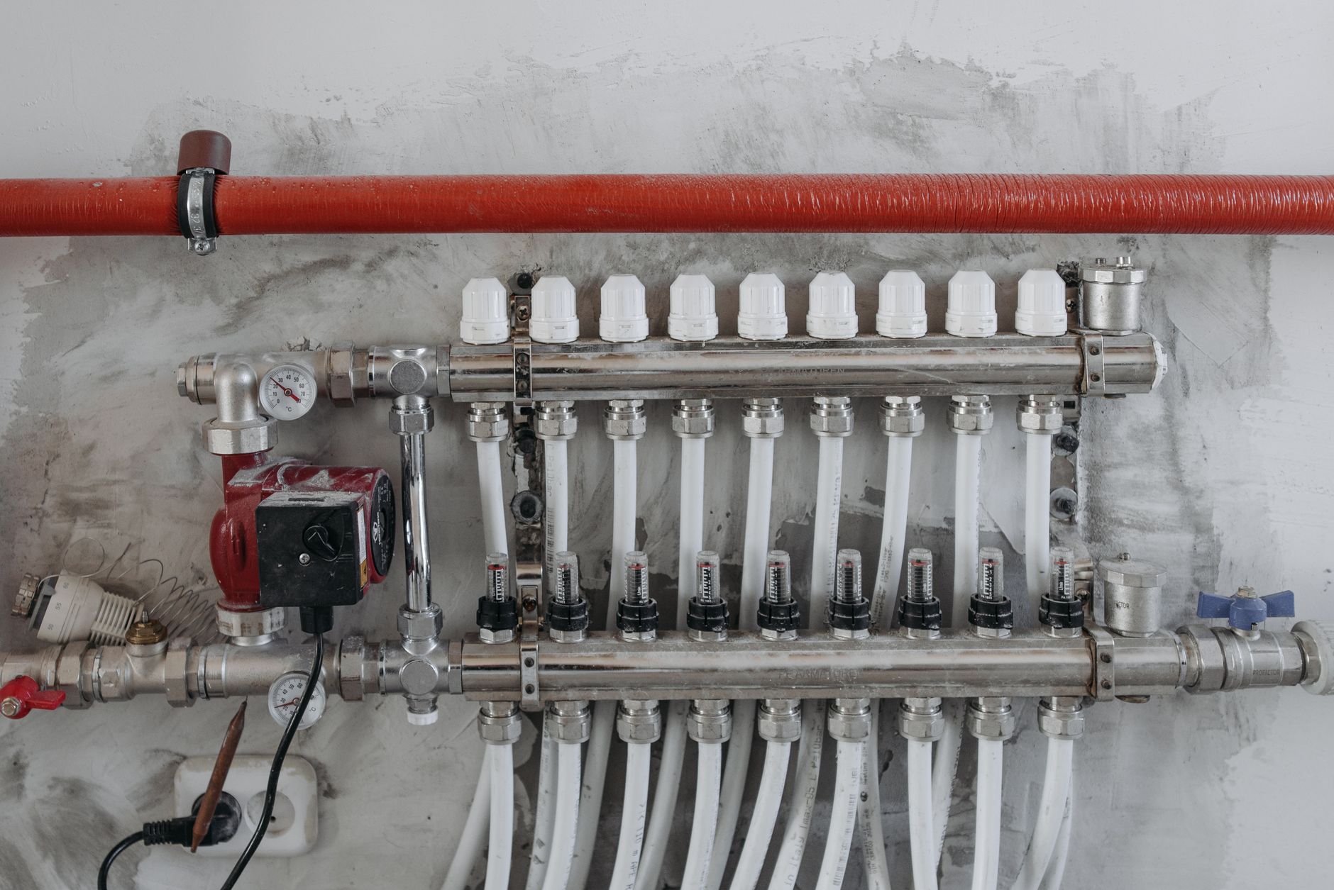 A hydronic heating manifold with white valve caps, flow meters, a red circulator pump and temperature gauges mounted on a concrete wall below a red insulated pipe