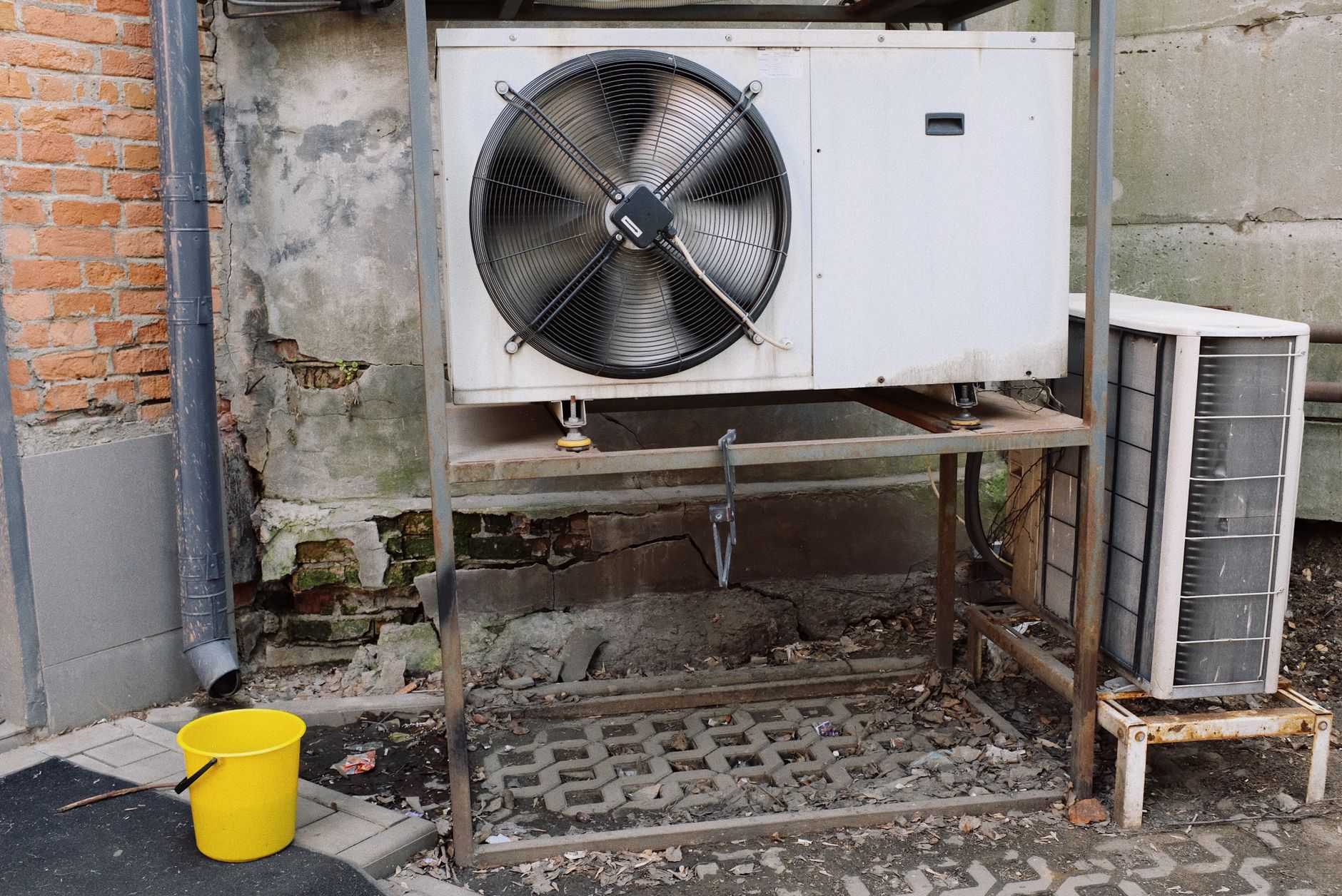 A commercial condensing unit with a large black fan on a steel stand beside a smaller split-system condenser against a brick and concrete wall