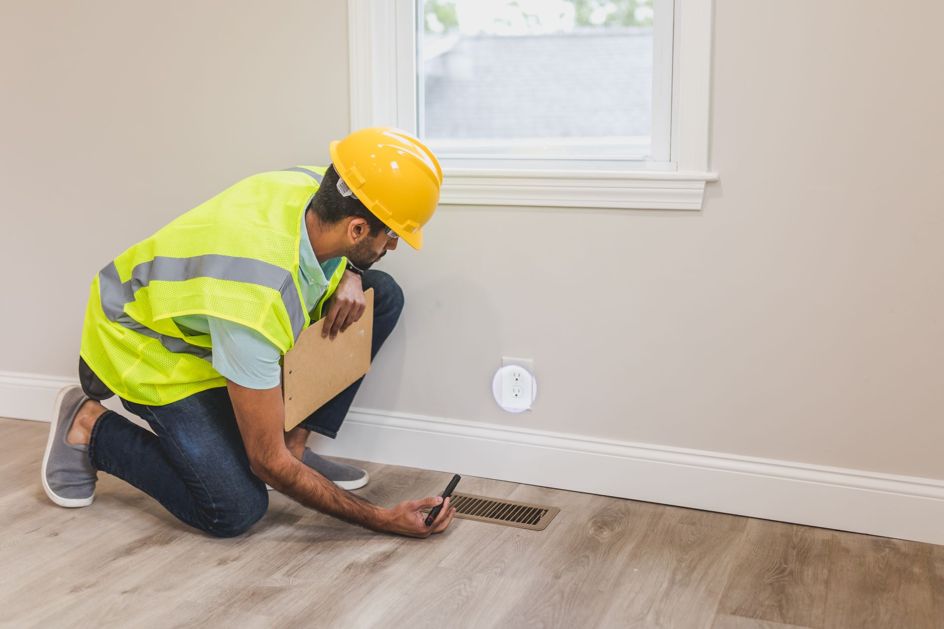A worker in a yellow hard hat and reflective vest kneels beside a floor supply vent, holding a clipboard and a small meter