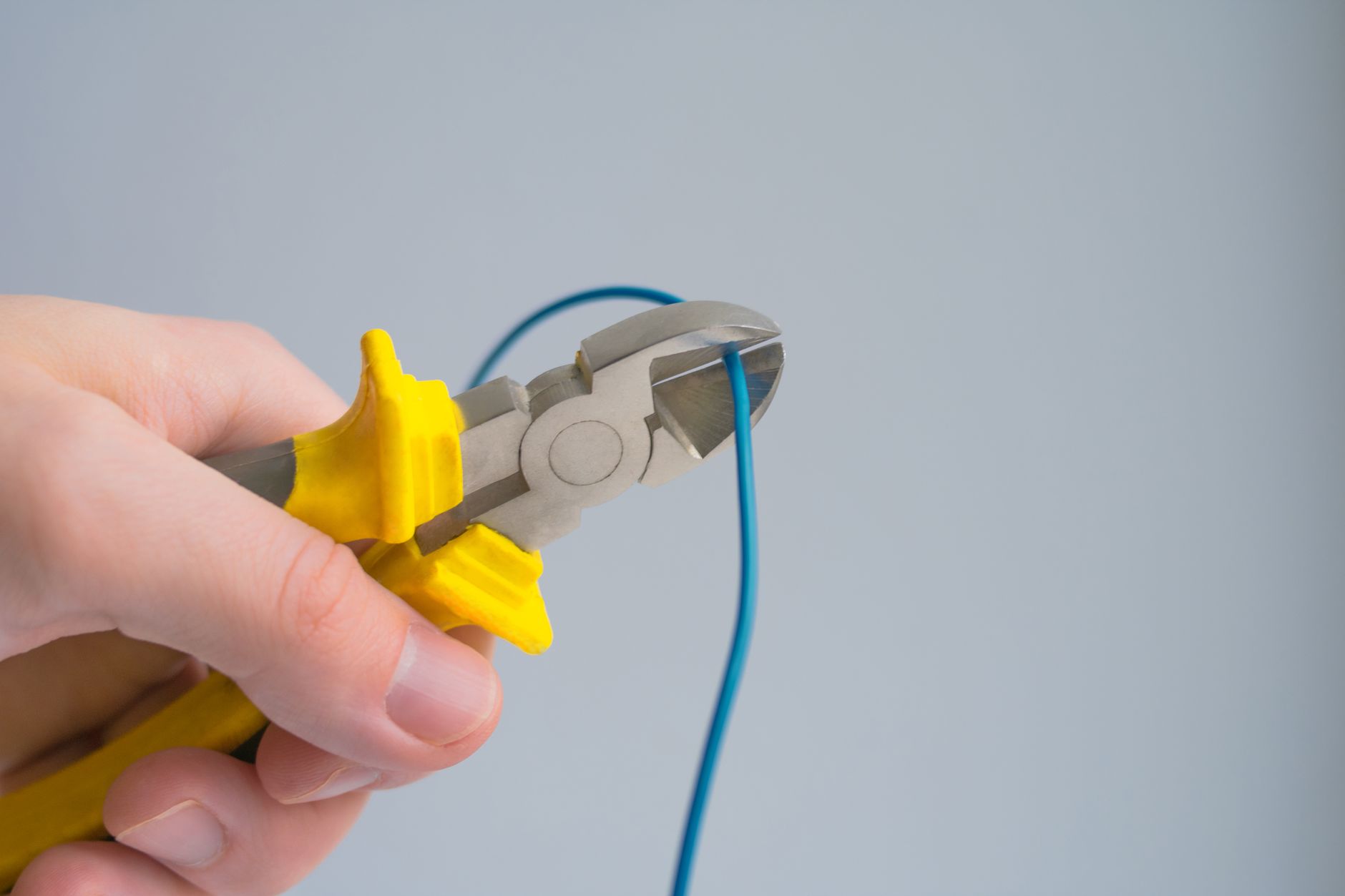 A hand holds yellow-handled diagonal cutters closed on a thin blue electrical wire against a plain gray background