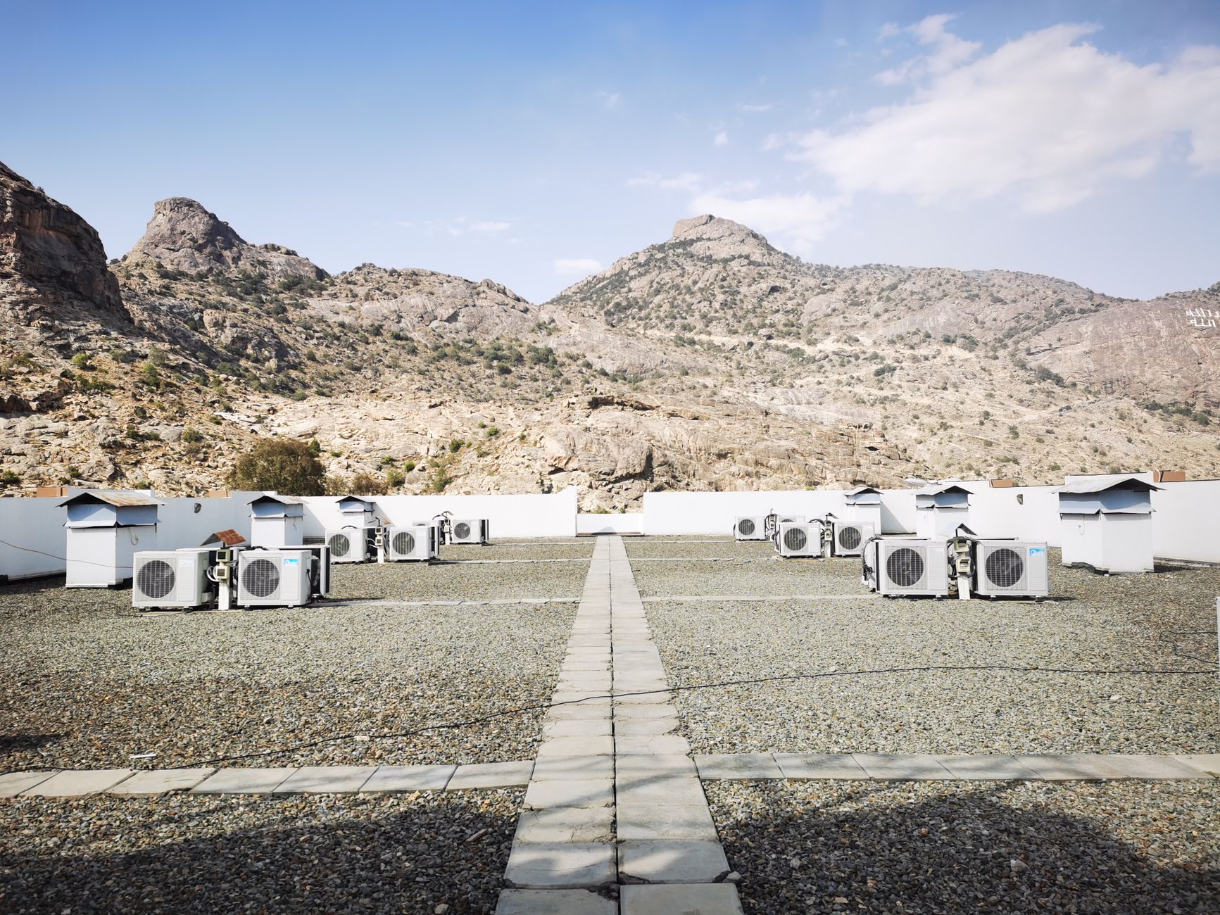 Rows of white air conditioning condenser units on a gravel rooftop with a paved walkway, rocky hills and blue sky behind