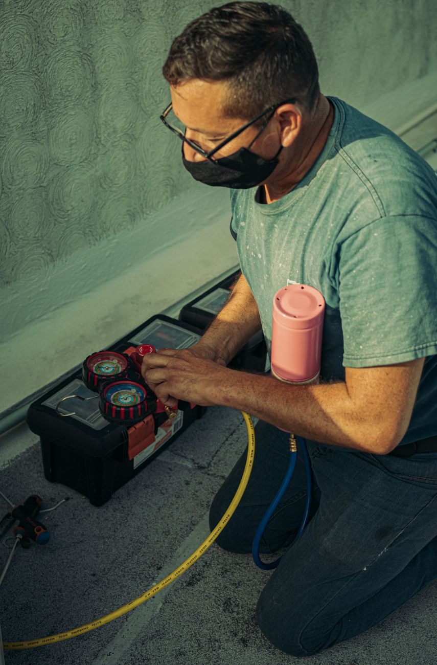 HVAC technician in glasses and a face mask kneeling on a rooftop to connect a red and blue manifold gauge set, with a pink refrigerant cylinder under his arm