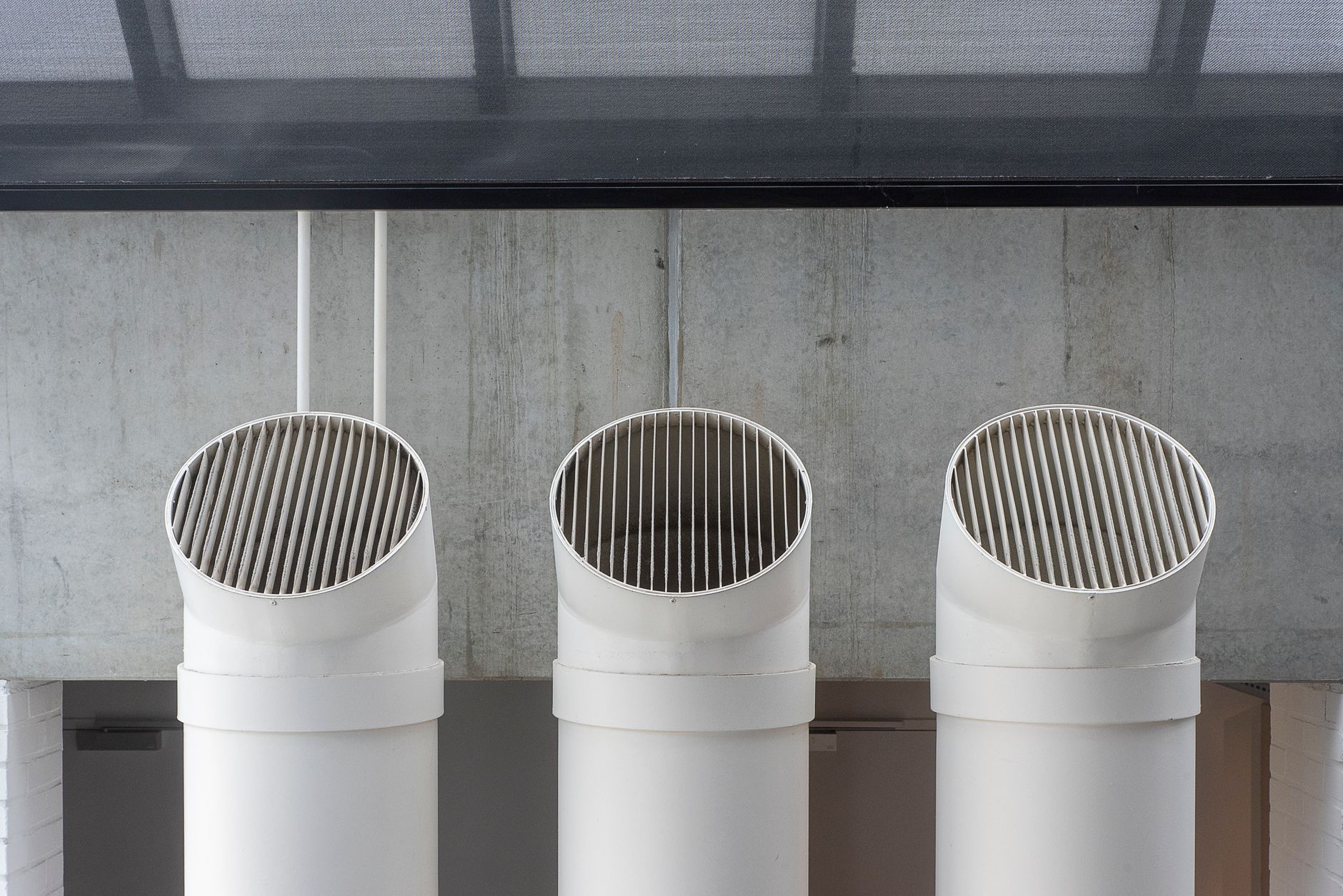 Three white round ventilation ducts with slatted grilles rise in front of a bare concrete wall under a dark ceiling