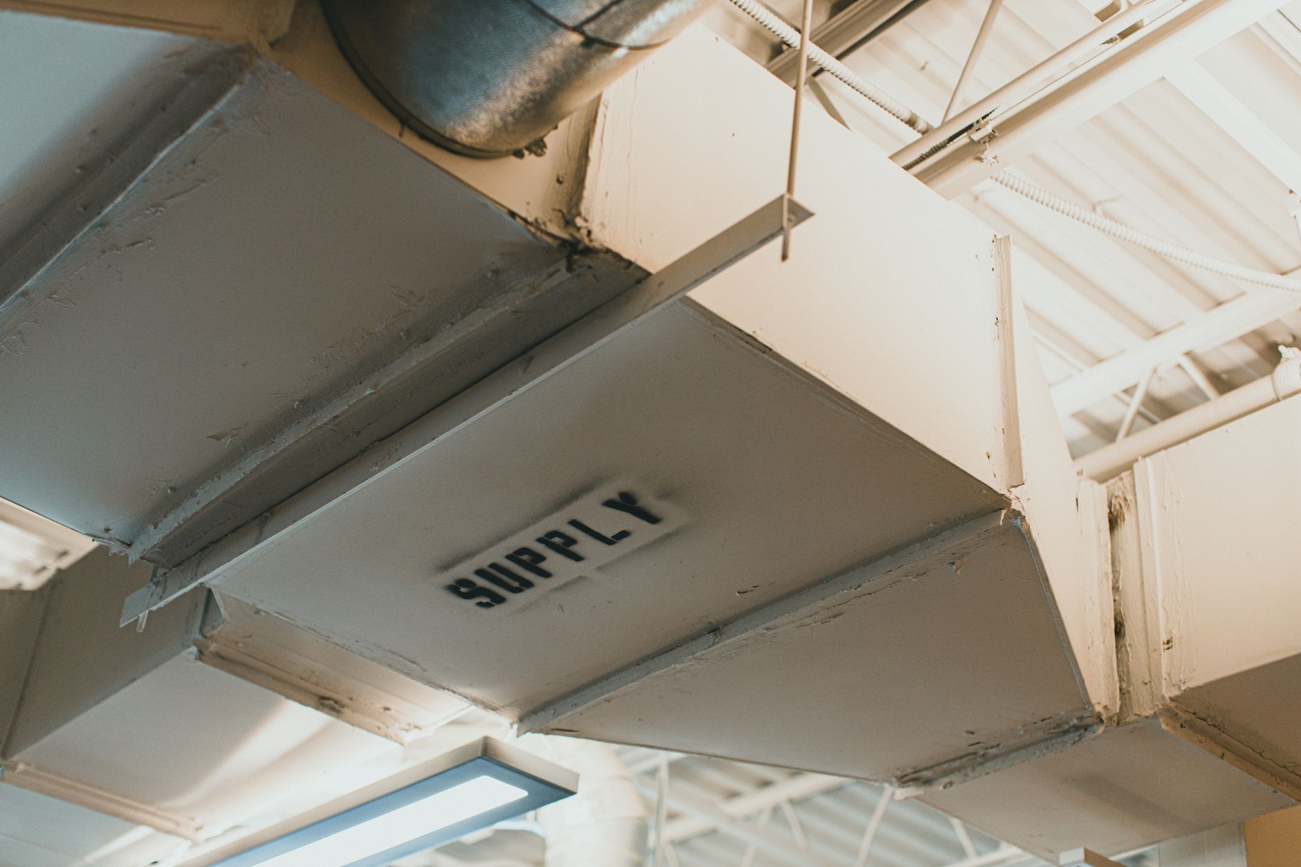 Painted rectangular sheet metal ductwork stenciled SUPPLY hangs from the ceiling of a commercial building beside a round duct