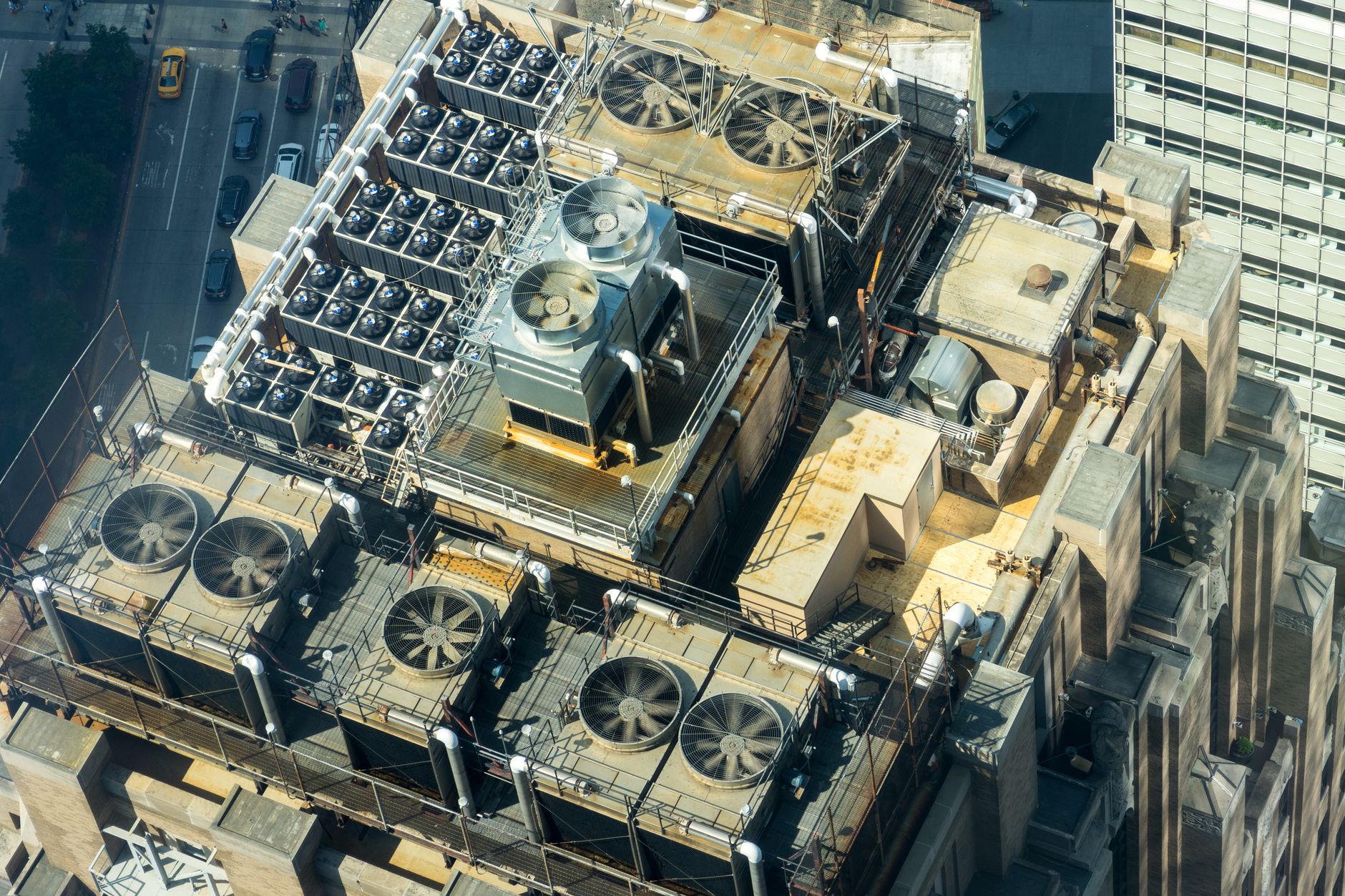 Aerial view of a high-rise rooftop packed with cooling towers, condenser fan banks, piping and catwalks, with a yellow taxi on the street below