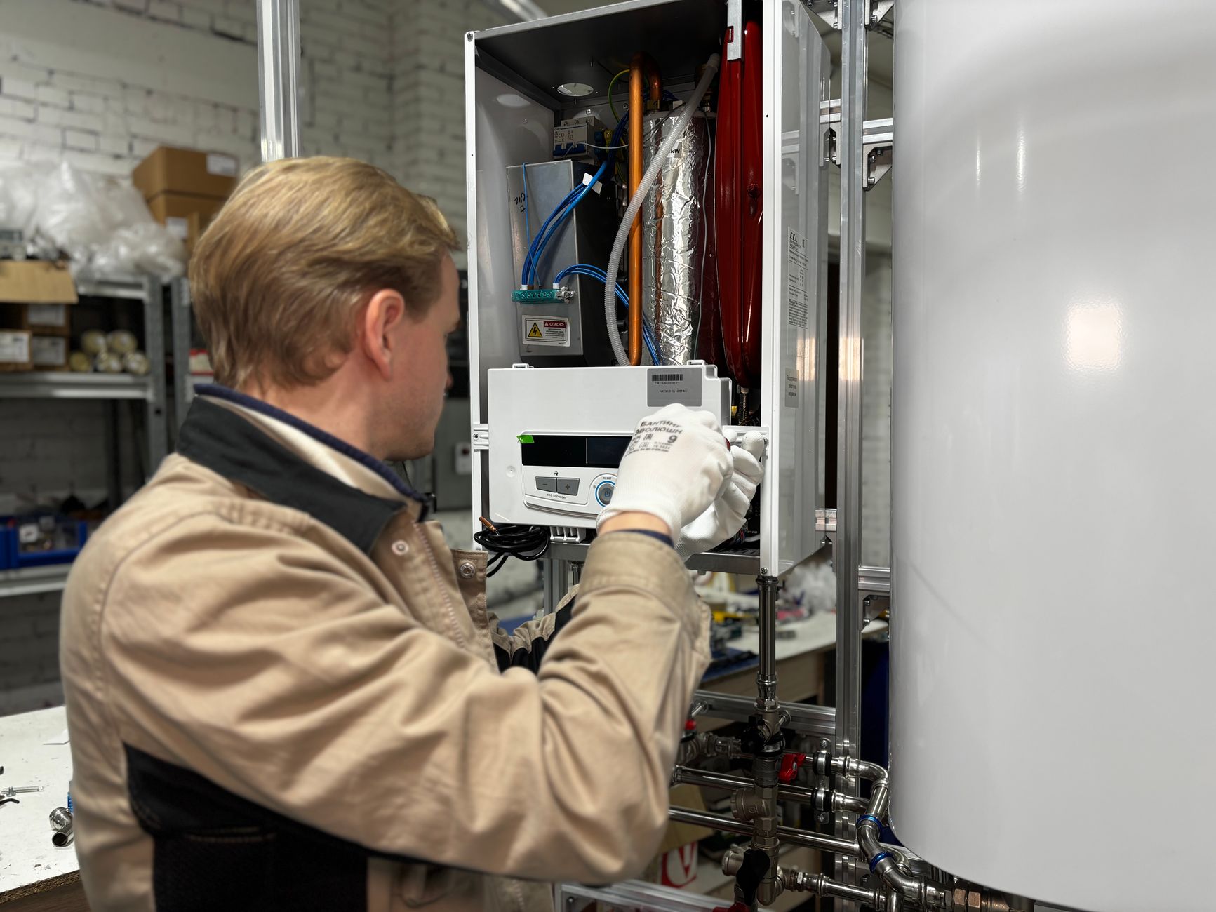 A technician in a tan work jacket and white gloves adjusts the controls of a wall-mounted heating unit with copper piping in a workshop