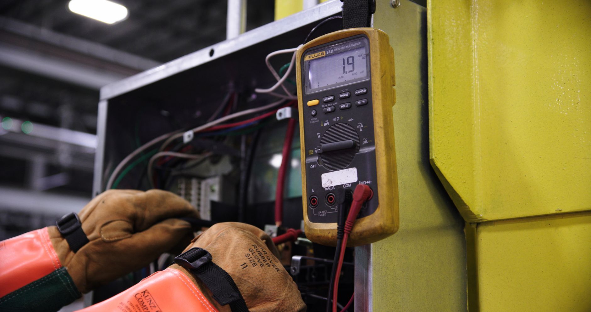 Gloved hands testing wiring inside an open equipment control panel with a yellow digital multimeter clipped beside it