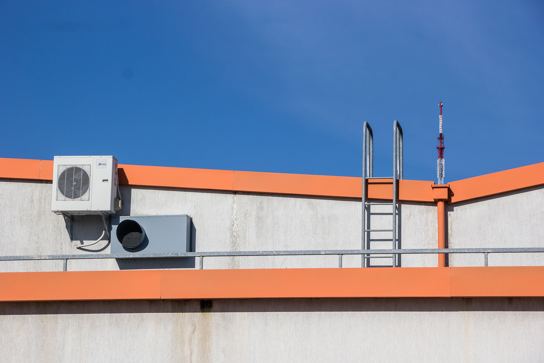 An outdoor air conditioning condenser mounted on a rooftop parapet next to a steel access ladder under a clear blue sky