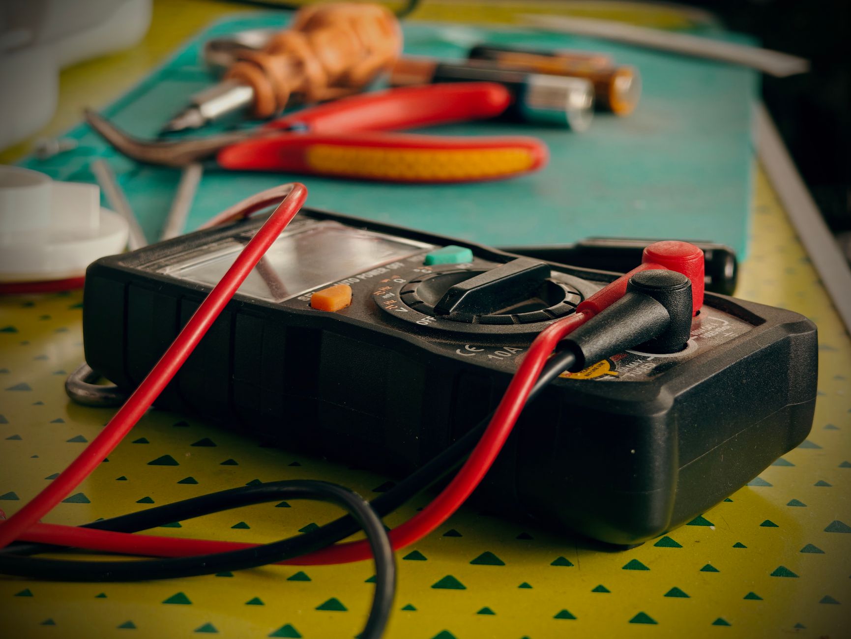 A black digital multimeter with red and black test leads on a workbench, with red-handled pliers and screwdrivers behind it