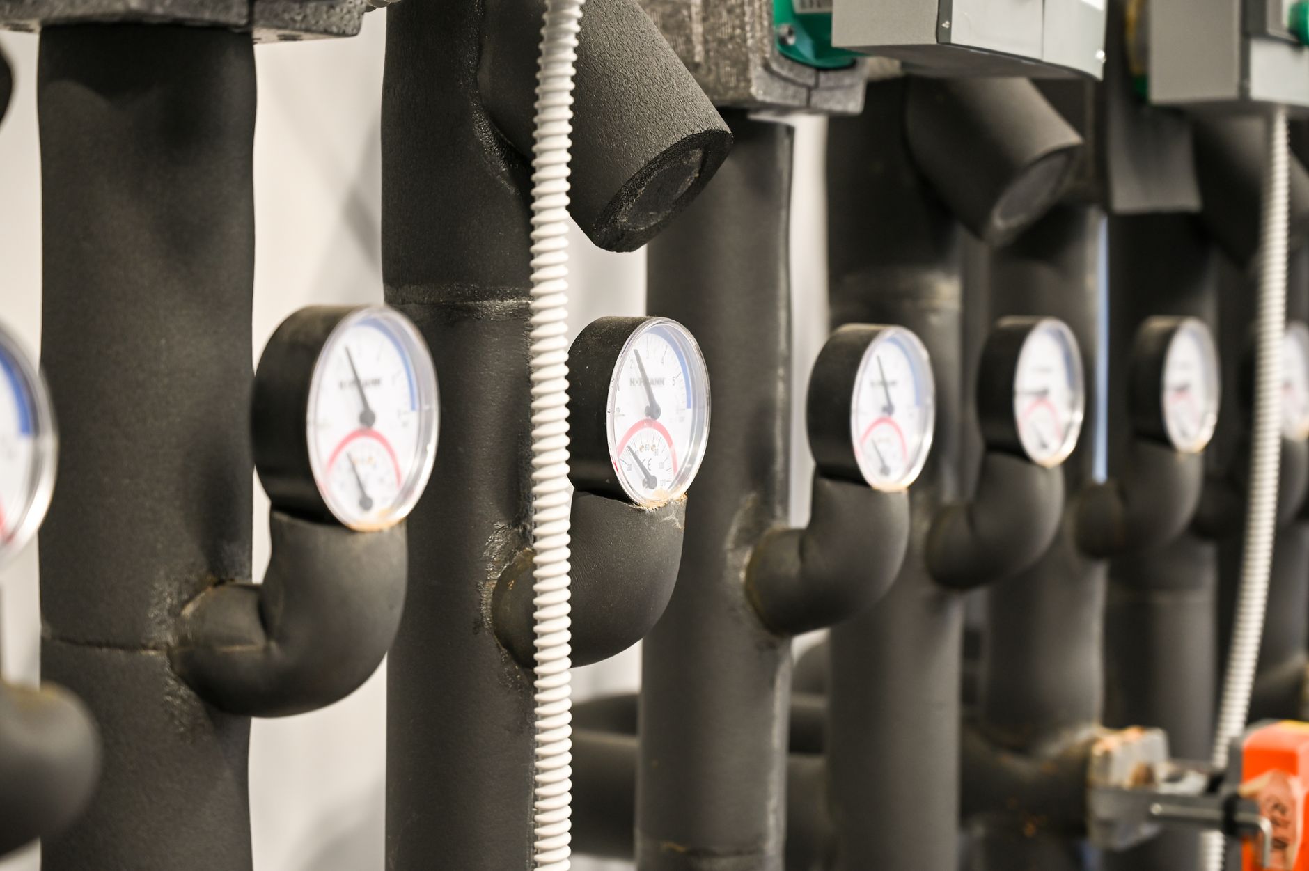 A row of insulated heating pipes, each fitted with a round temperature and pressure gauge, in a mechanical room