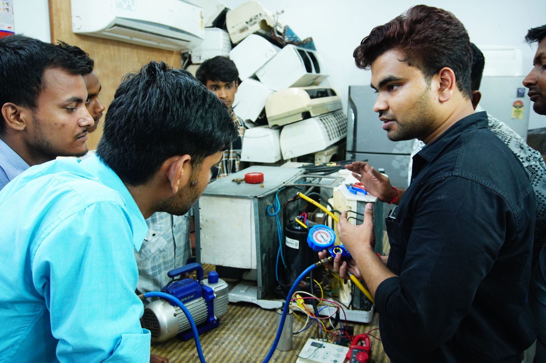 An instructor showing students a manifold gauge beside a vacuum pump and an open air conditioning trainer