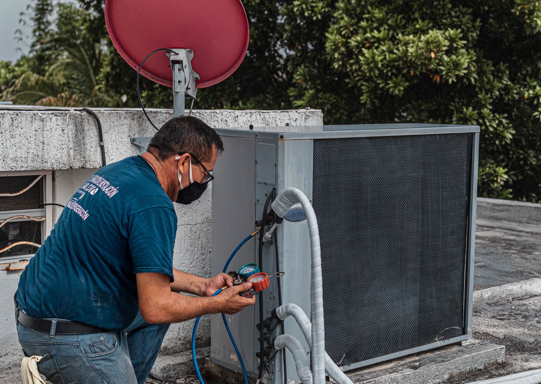 An HVAC technician kneeling at a rooftop condenser, reading a manifold gauge set connected by refrigerant hoses